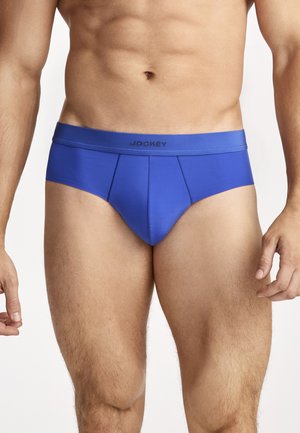 Männlicher Torso mit hellblauer Slip-Unterwäsche und Jockey-Logo am Bund, vor einfachem weißem Hintergrund stehend.