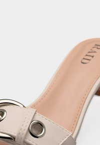 Mule en cuir beige avec détail de boucle argentée et semelle intérieure cousue, présenté en angle pour montrer la semelle et une partie du talon.