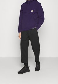 Lilla pulloverjakke med hætte, frontlomme og snor. Sammen med løstsiddende sorte bukser og sorte sneakers. Enkel, funktionel design.