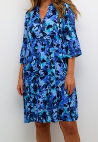 Kvinna i en knälång blå blommig klänning med 3/4 ärmar och V-ringning mot en enkel vit bakgrund.