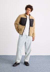Fleecejacke in Beige mit schwarzem Nylon-Brustpanel, Reißverschlusstasche, Stehkragen, kombiniert mit heller Jeans und schwarzen Schuhen mit roten Akzenten.