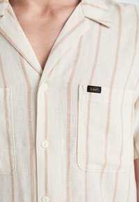 Light-colored short-sleeve shirt with vertical beige stripes, a patch pocket, and a black "Lee" label on the chest. Buttoned front.