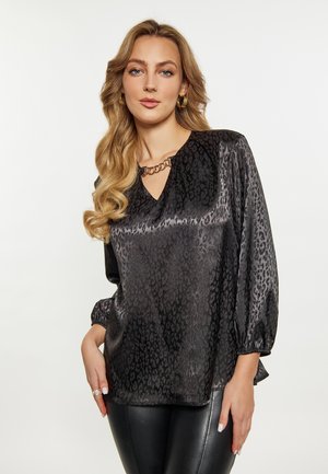 Femeie cu păr blond lung poartă o bluză din satin negru cu model și detaliu cu lanț și pantaloni de piele negri pe un fundal simplu și luminos.