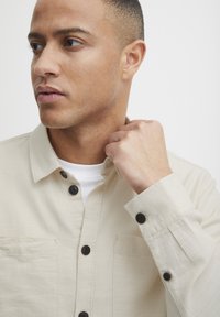 Camisa de botón beige con cuello, botones negros y dos bolsillos en el pecho. La tela parece ser texturizada y ligera.