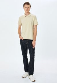 Beige polo shirt med lynlåskrave, parret med mørkeblå denimjeans og hvide sneakers. Stoffet ser glat og letvægts ud.
