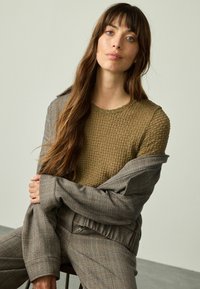 Olivgrüner, strukturierter Pullover unter einem grauen, karierten Oversized-Blazer; Modell mit langen Haaren, sitzend, mit übergeschlagenen Armen, neutraler Hintergrund.