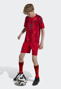 Punainen Bayern München -paita, jossa mustat yksityiskohdat, sävyyn sopivat punaiset shortsit ja punaiset sukat. Pelaaja seisoo mustan ja valkoisen jalkapallon kanssa.