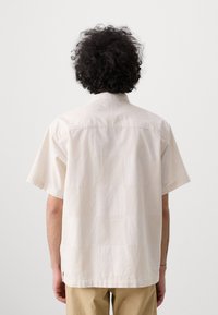 Chemise à manches courtes, couleur écru, fabriquée en tissu texturé, avec un empiècement au dos et des fentes sur les côtés ; design décontracté et relaxé.