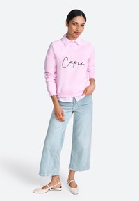 Felpa rosa con ricamo "Capri", indossata sopra una camicia bianca con colletto, abbinata a jeans a gamba larga blu chiaro e scarpe slingback chiare.