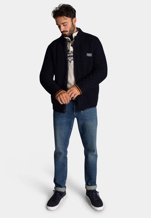 Marineblauwe gebreide cardigan met een textuurpatroon, oranje accentmanchetten, over een lichte trui, gecombineerd met blauwe jeans en donkere sneakers.