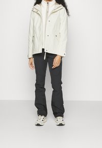 Chaqueta crema ligera con capucha, cremallera frontal y cierre de botón a presión, combinada con pantalones negros impermeables y zapatillas beige.