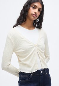 Κρεμ ριμπ ζακέτα με βαθύ V-λαιμό, κουμπιά μπροστά και κυματιστό τελείωμα. Φορεμένη πάνω από λευκό τοπ, συνδυασμένη με σκούρο τζιν.