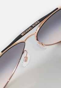 Roségoldene Aviator-Sonnenbrille mit graduellen grauen Gläsern, schwarzen Bügelakzenten und "MICHAEL KORS"-Branding an der Oberseite des Gestells.
