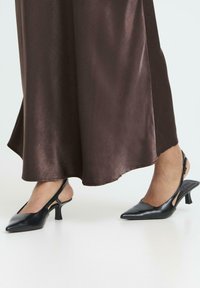 Juodos odinės slingback kulniukai su smailiu priekio ir mažu kubiniu kulnu, derinami su plazdančia rudą satino sijonu.