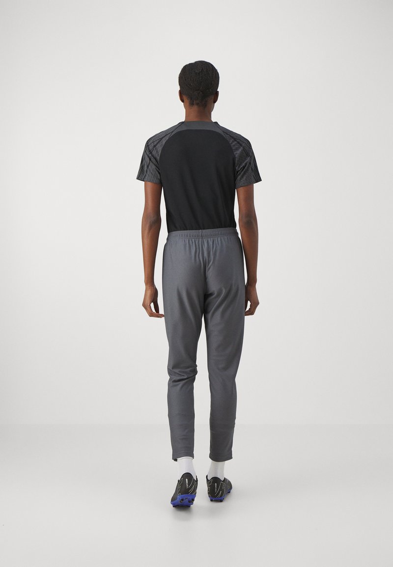 Persona con una camiseta de manga corta negra y gris, pantalones grises, calcetines blancos y botas negras con suelas azules, de pie de espaldas sobre un fondo blanco.