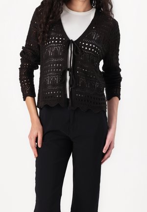 Woman wearing a black open-knit cardigan tied at two points over a white ribbed top and black pants, standing with hands by sides.