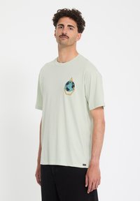 Mann mit lockigem Haar und Schnurrbart, der ein hellgraues T-Shirt mit einer blauen und gelben Globusgrafik auf der Brust und eine schwarze Hose trägt.