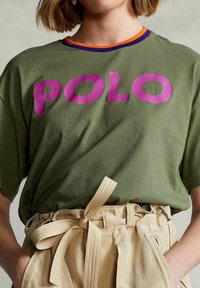 Olive green cotton T-shirt with large purple "POLO" print, contrasting orange and purple neckline. Paired with beige high-waisted trousers.