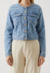 Vrouw in een lichtblauwe denim jas met knopen en borstzakken, in combinatie met beige high-waist broek, staat tegen een effen achtergrond.