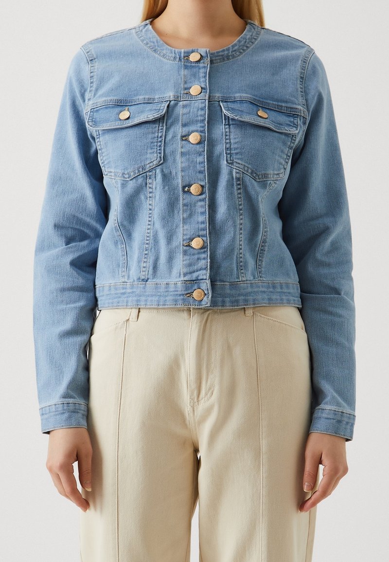 Vrouw in een lichtblauwe denim jas met knopen en borstzakken, in combinatie met beige high-waist broek, staat tegen een effen achtergrond.