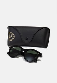 Melnas Ray-Ban saulesbrilles ar zaļām lēcas, kurām ir kaķa acs forma un izteiksmīgi tempļi. Komplektā ar teksturētu melnu futlāri.