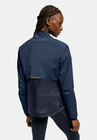 Marineblaue Sportjacke mit verkürztem Rücken und glänzender Textur, ausgestattet mit einem hohen Kragen und einem Logo-Akzent auf der linken unteren Seite.