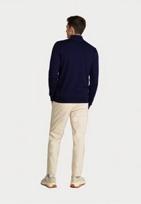 Junger Mann, der mit dem Rücken zur Kamera steht, trägt einen navyblauen Pullover, beigefarbene Hosen und weiße Sneakers vor einem schlichten, hellen Hintergrund.