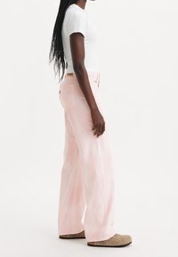 Personne avec de longs cheveux tressés portant un t-shirt blanc, un pantalon large rose pâle et des chaussures beige à enfiler, se tenant de profil.