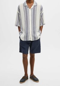 Camicia a righe a maniche corte in beige e blu navy, con chiusura a bottoni. Abbinata a pantaloni corti blu scuro e scarpe scure con lacci marroni.