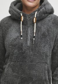 Grijze fleece hoodie met een ritssluiting aan de voorkant, trekkoordsluiting en een geweven stof. Voorzien van houten accenten en contrasterende koorden.