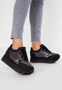 Mustat ja hopeiset korkeat tennarit, joissa on teksturoitu glitterpinta, pyöreä varvas ja tyylikäs musta kumipohja, jossa on vaakaraitoja.