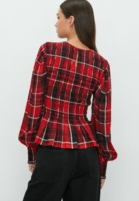 Blouse à carreaux rouges avec de longues manches bouffantes, un panneau arrière smocké et un ourlet peplum. Présente des détails blancs et noirs dans le motif.