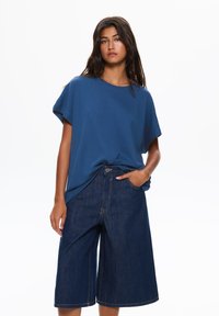 T-shirt blu dalla vestibilità ampia con maniche corte, abbinato a culotte in denim blu navy con tasca frontale e dettagli di cucitura visibili.