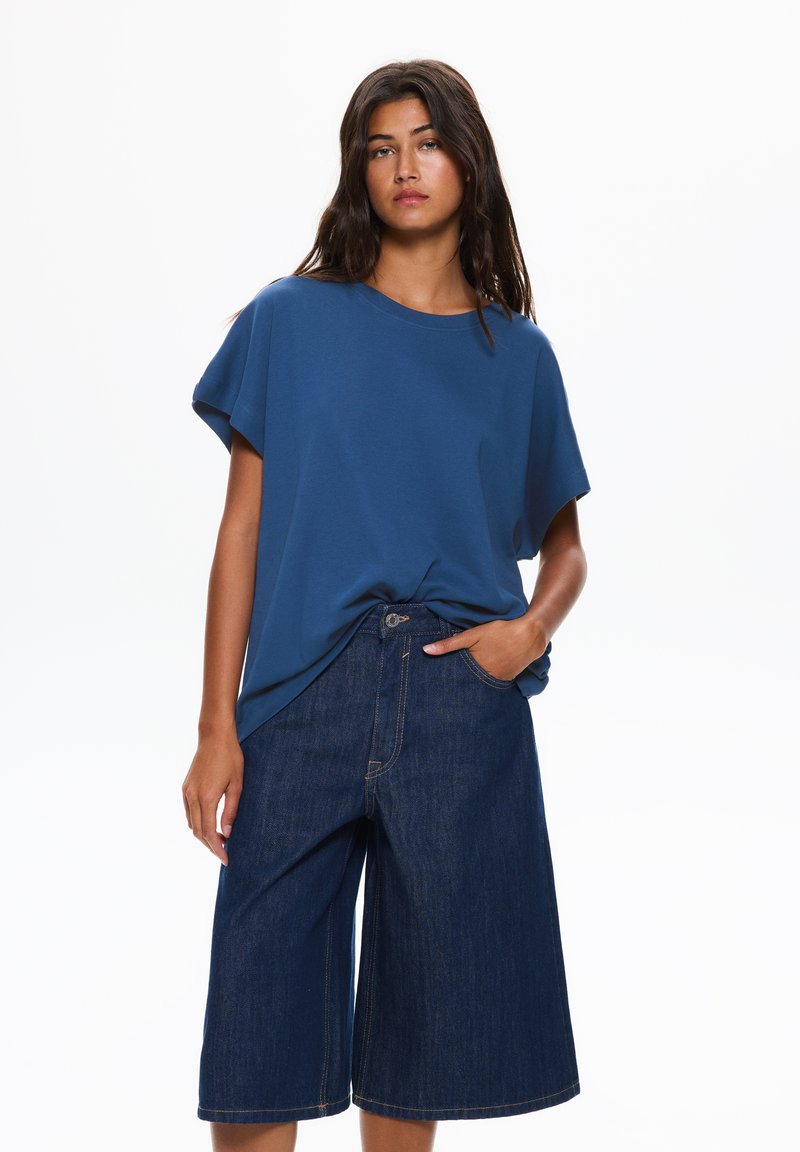 T-shirt blu dalla vestibilità ampia con maniche corte, abbinato a culotte in denim blu navy con tasca frontale e dettagli di cucitura visibili.
