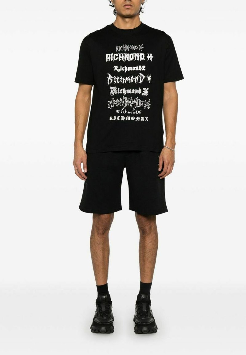 T-shirt in cotone nero con audaci design testuali bianchi disposti verticalmente. Abbinato a pantaloni corti neri e sneakers nere di grandi dimensioni.