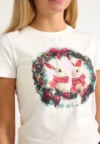 Hvid bomulds t-shirt med et grafisk motiv af to kaniner i en festlig krans, prydet med grønne blade, røde sløjfer og dekorative bær.