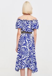 Osoba z blond włosami widziana z tyłu, ubrana w niebieską i białą sukienkę bez ramiączek w zebrę oraz z wcięciem w talii.