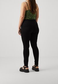 Kobieta ubrana w czarne dopasowane spodnie, zielono-czarny top bez rękawów z wzorem oraz czarne sandały na platformie, stojąca na tle jednolitego szarego tła.