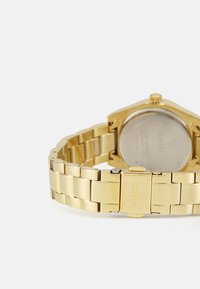 Goldfarbene Metall-Armbanduhr mit einem Gliederarmband, runder Gehäuseform und einem Gehäuseboden aus rostfreiem Stahl. Verfügt über eine Schließe mit eingraviertem "Guess".