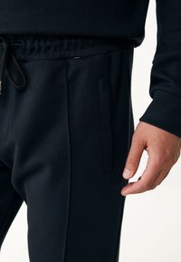 Pantalones de chándal negros con cintura ajustable, bolsillos laterales y un diseño ajustado. La tela parece suave y tiene una textura lisa.