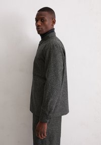 Grau, strukturierte Jacke mit Kragen, Knopfmanschetten und einer Brusttasche, kombiniert mit einem passenden dunklen Outfit.