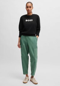 Black sweatshirt with "BOSS" printed in white, paired with loose green twill pants. Shoes are light beige with a textured design.