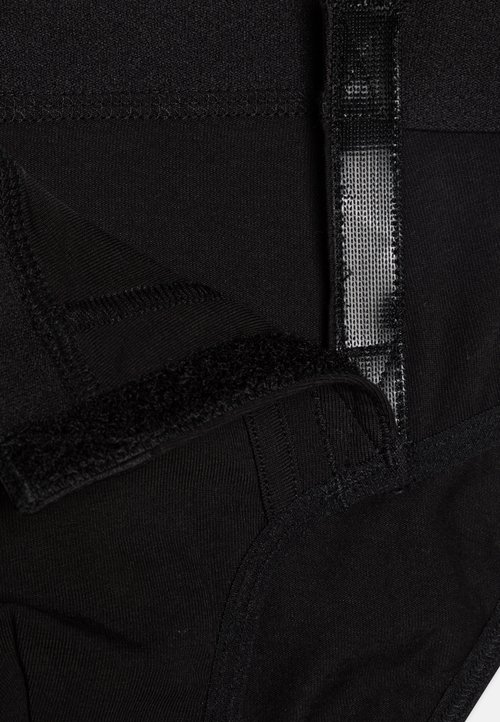 Juodas audinys drabužis, turintis tekstūruotą paviršių, siuvinėtus kraštus ir Velcro užsegimą. Dizainas yra lygus, su paslėptu vidiniu sluoksniu.