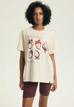 Kobieta ubrana w kremowy T-shirt z czerwono-brązowymi ilustracjami truskawek i napisem "SUMMER MARKET", w połączeniu z bordowymi szortami.