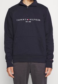 Navy blue sweatshirt with a high collar and quarter zipper, features a front pocket and embroidered "TOMMY HILFIGER EST. 1985" logo.