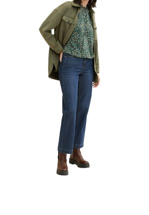 Grünes übergroßes Hemd-Jackett, Bluse mit Blumenmuster, blaue gerade Jeans und braune Knöchelstiefel mit dicken Sohlen.