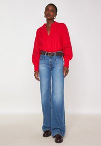 Camicia rossa a maniche lunghe con colletto a bottoni e dettagli in occhiello, abbinata a jeans blu a gamba larga e una cintura marrone, indossata con scarpe nere.
