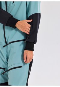 Een tweekleurige jumpsuit met een teal bovenstuk en zwarte ondermouwen, voorzien van ritszakken en ribgebreide manchetten.