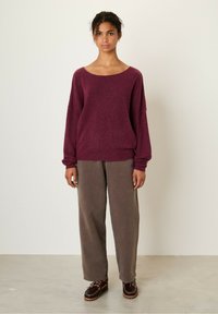 Bordeaux gebreide trui met een brede halslijn, oversized pasvorm en ribbels aan de mouwen, gecombineerd met losse grijze broek. Bruine veterschoenen maken de look compleet.