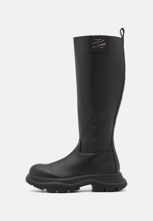 Botte noire montant jusqu'au genou avec semelle épaisse texturée, bout arrondi et languette de tirage en haut sur un fond clair uni.